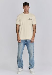 Camiseta de algodón beige con mangas cortas, que presenta un logo en cursiva negro en el pecho. Combinada con jeans azules descoloridos y zapatillas blancas.