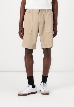 Beige cargo shorts made of cotton blend fabric. Features two side pockets and a relaxed fit. Paired with white sneakers and black socks.