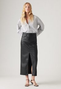 Jupe midi noire en simili cuir avec une fente avant, associée à une chemise à rayures bleues et blanches à boutons, et des talons noirs à lanières.