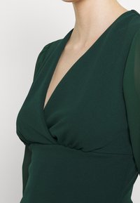 Tmavě zelené šaty s hlubokým véčkovým výstřihem, nařaseným korpusem a průhlednými dlouhými rukávy. Látka je hladká s mírným elastickým efektem.