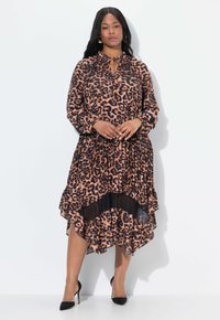 Donna che indossa un vestito a maniche lunghe con stampa leopardata marrone e nera, con orlo plissettato e tacchi alti neri, in piedi su uno sfondo bianco.