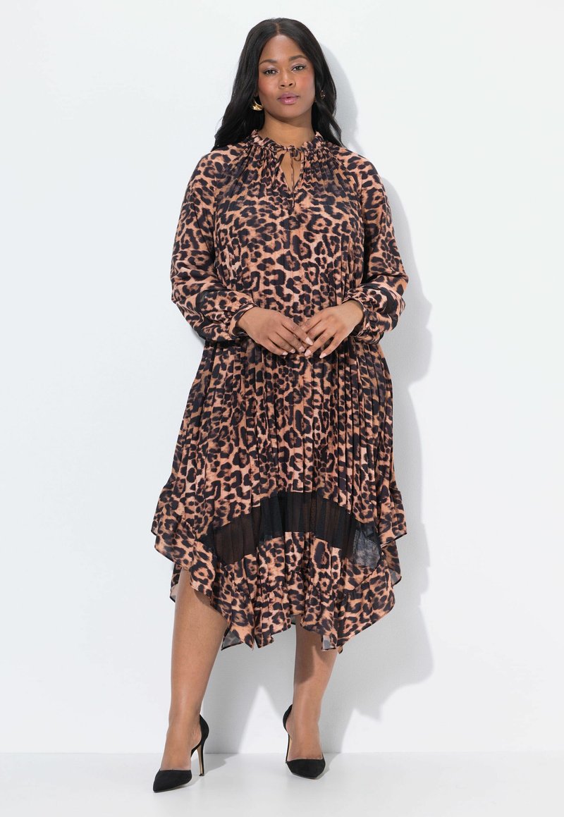 Donna che indossa un vestito a maniche lunghe con stampa leopardata marrone e nera, con orlo plissettato e tacchi alti neri, in piedi su uno sfondo bianco.