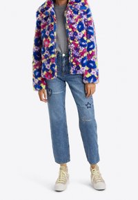 Kolorowa kurtka z imitacji futra zapinana na zamek, z kwiatowym wzorem w różu, niebieskim i żółtym, zestawiona z jasnoniebieskimi dżinsami i beżowymi trampkami.