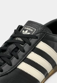 Μαύρο δερμάτινο αθλητικό παπούτσι με τρεις κρεμ ρίγες, κρεμαστό μαύρο γλώσσον και μαύρα κορδόνια. Διαθέτει ανάγλυφο λογότυπο στη γλώσσα.