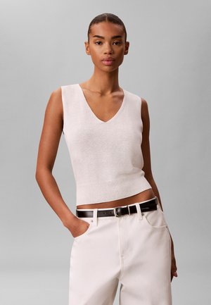 Femme mince aux cheveux tirés en arrière portant un haut blanc sans manches à col en V, un pantalon blanc et une ceinture noire, debout avec une main dans la poche.