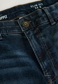 Slim fit dark blue denim jeans with a metallic button, visible stitching, and a label reading "Supply" and size "34 R."