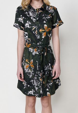 Kvinde iført kortærmet knap-op kjole med grønne blade og orange blomsterprint, bundet ved taljen, stående mod en ensfarvet baggrund.