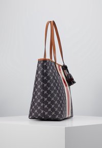 Plátěná taška s tmavě modrým základem zdobeným bílým vzorem listů, hnědými koženými popruhy a dekorativním třásňovým akcentem. Na boku jsou svislé červené pruhy.