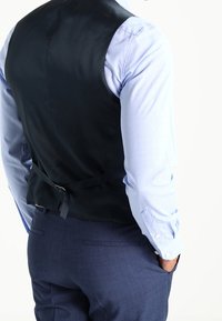 Giubbotto in satin nero con una texture liscia, design aderente e cinturino posteriore regolabile, indossato sopra una camicia azzurra a motivo e pantaloni blu navy.