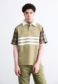 Khaki polo majica z belim ovratnikom, z značilnimi karirastimi rokavi in tremi belimi črtami čez prsni del; v kombinaciji z ustreznimi olivno zelenimi hlačami.