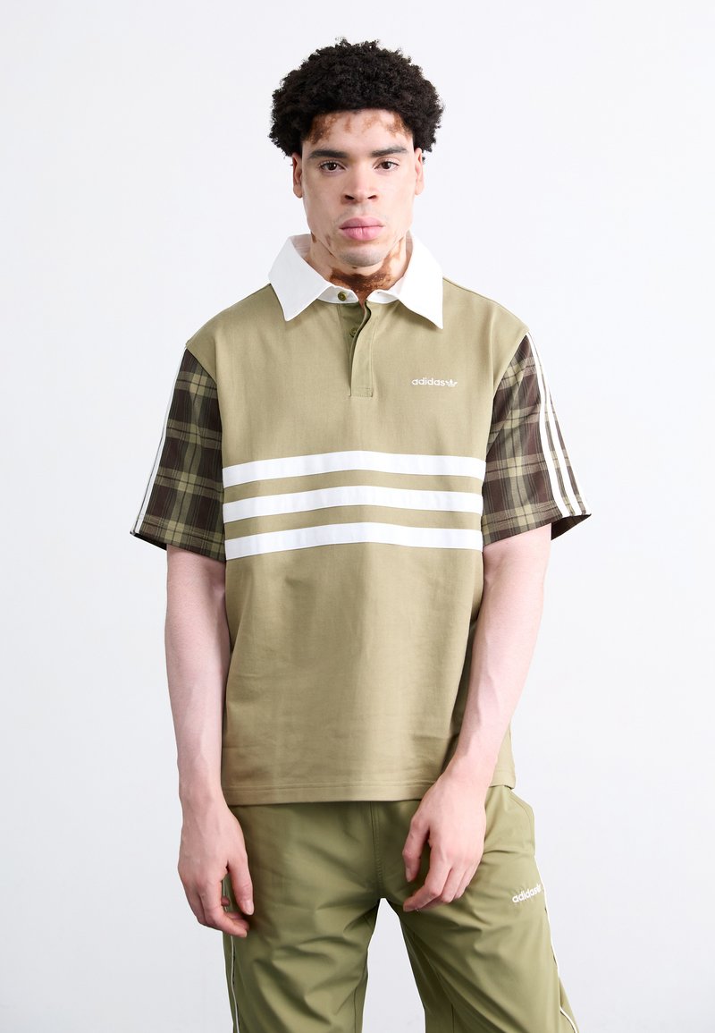 Khaki polo majica z belim ovratnikom, z značilnimi karirastimi rokavi in tremi belimi črtami čez prsni del; v kombinaciji z ustreznimi olivno zelenimi hlačami.