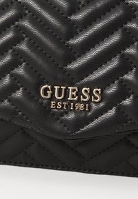 Schwarze, gesteppte Lederhandtasche mit einem goldfarbenen "GUESS EST 1981"-Logo. Strukturiert Oberfläche mit ausgeprägten Nähten und Fischgrätmuster.