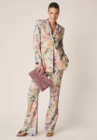 Frau trägt einen pastellfarbenen Blumenanzug mit passenden weit geschnittenen Hosen, hält eine mauvefarbene Wildleder-Clutch und steht mit einer Hand in der Hosentasche.