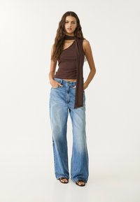 Top marrone senza maniche con dettaglio di sciarpa e jeans a gamba larga blu. I jeans sono strappati con orlo leggermente sfilacciato. Sandali neri completano il look.
