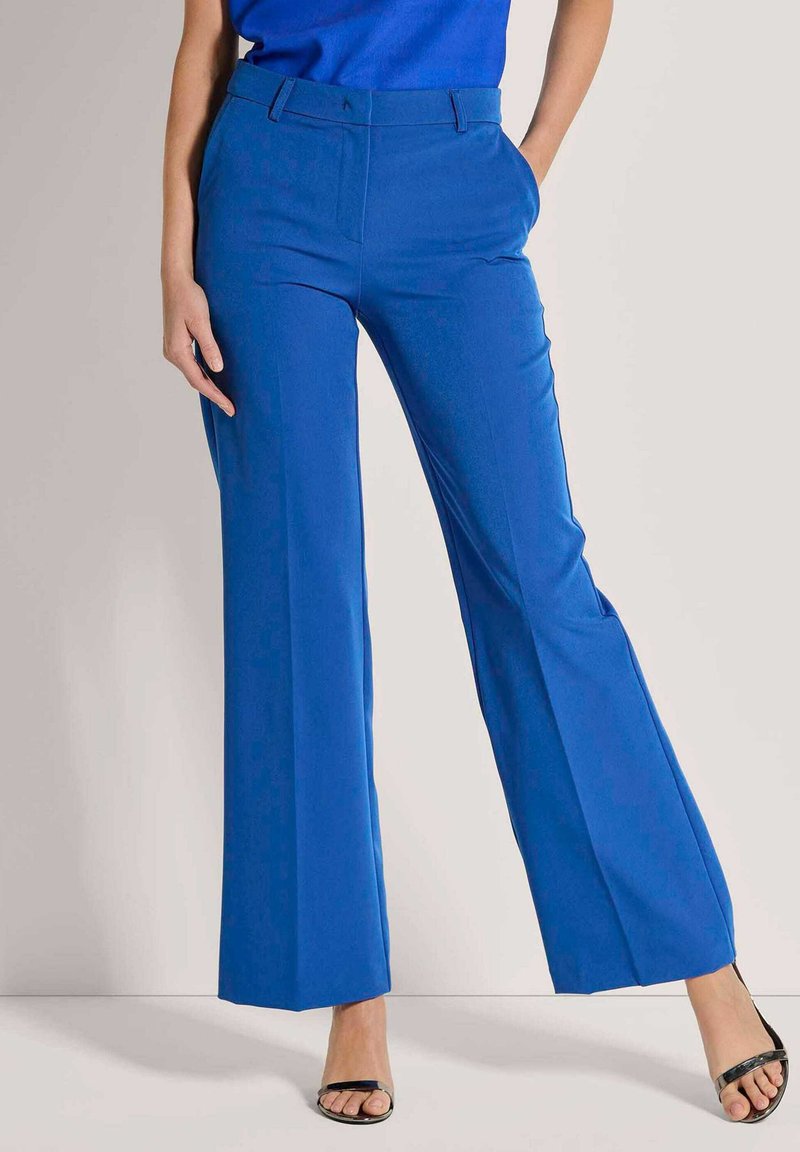 Blauwe op maat gemaakte wijde broek van een gladde stof. Heeft een vlakke voorkant, riemlussen en zijzakken. G gedragen met eenvoudige zwarte sandalen.