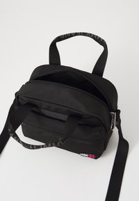 Sac en tissu noir avec deux anses supérieures, une bandoulière amovible, des fermetures éclair doubles et un patch logo. Forme compacte et rectangulaire avec une surface texturée.