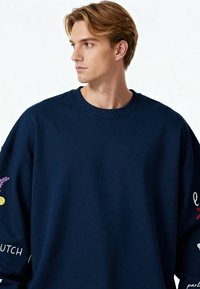 Mornarsko modra oversize majica z dolgimi rokavi z rebrastim ovratnikom in dekorativnim vezenjem na rokavih, vključno s cvetličnimi vzorci in besednimi poudarki.