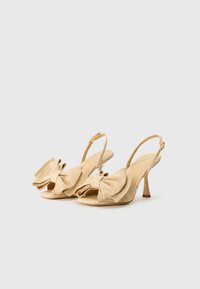 Šviesiai rudos slingback aukštakulniai sandalai, pasižymintys dideliu kaspinu priekyje, atviru pirštu ir tekstūruota medžiaga. Kulnas yra siauras ir maždaug 5 cm aukščio.