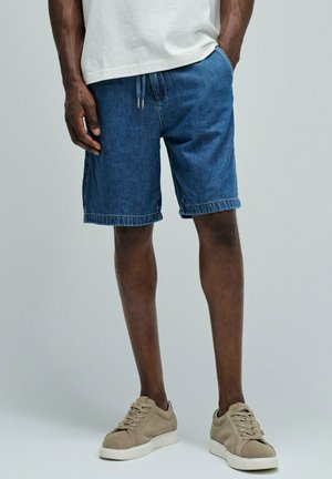 Hombre con pantalones cortos vaqueros azules, camiseta blanca y zapatillas beige, de pie con una mano en el bolsillo contra un fondo liso.