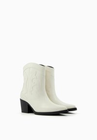 Białe kowbojskie buty o gładkim wykończeniu ze skóry, z delikatnym falistym haftem, spiczastym noskiem i solidnym czarnym obcasem w kształcie bloku.