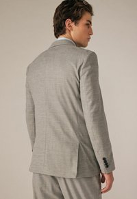 Hellgraue Jacke mit Karo-Muster, strukturierten Schultern und einem Schlitz. Schwarze Knöpfe an den Ärmelabschlüssen setzen Akzente.