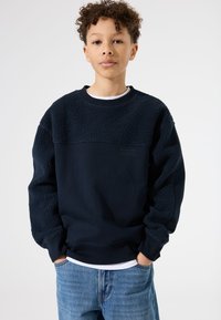 Sudadera azul marino con hombros y mangas de textura tipo forro polar. Presenta un cuello redondo y puños acanalados. Combinada con jeans de color azul claro.