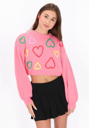 Pulover roz scurt cu modele de inimi multicolore, guler ondulat și mâneci tip balon, asortat cu o fustă neagră plisată.