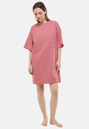 Femme pieds nus portant une robe rouge ample longueur genou avec un motif répétitif de cœurs, debout avec une posture détendue devant un fond blanc uni.