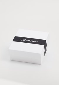 Baltas stačiakampis langelis su lygiu paviršiumi, su juodu juostele, pažymėta „Calvin Klein“ ryškiu bešriftu šriftu.
