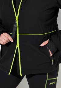 Osoba v černé bundě s neonově žlutým zipem a lemováním, ruce u zipu a kapsy, v kombinaci s černými legínami s neonovým prošitím.