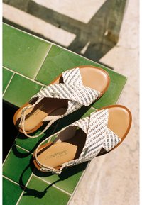 Woven sandals with a crisscross design in beige and white, featuring a brown sole and an adjustable ankle strap. Decorative logo on the insole.