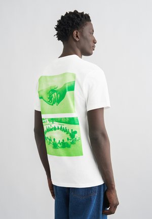 Egy férfi fehér, zöld nyomtatású pólóban, amelyen két kéz fogja egymást, és egy csoport ember ül kint, körben.