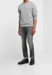 Sudadera gris con el logo "HACKETT", combinada con jeans gris oscuro remangados en los tobillos y zapato negro pulido tipo oxford. Diseño minimalista.