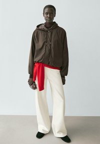 Frau trägt eine braune Kapuzenjacke, roten Pullover um die Taille gebunden, weiße Hosen mit weitem Bein, schwarze Schuhe, hält eine kleine braune Tasche.