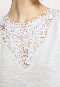 Vit kortärmad topp med intrikata spetsdetaljer runt halslinjen, vilket ger textur och designkontrast till det släta tyget.