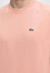 Άτομο που φοράει ανοιχτό ροζ μπλουζάκι με στρογγυλή λαιμόκοψη και μικρό πράσινο λογότυπο κροκόδειλου κεντημένο στο αριστερό μέρος του στήθους.