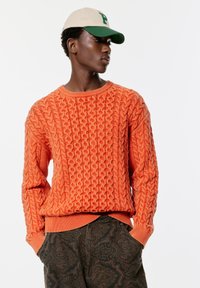 Orange stickad tröja med ett upphöjt kabelmönster, ribbad rund halsringning och ärmslut. Matchad med mörka mönstrade byxor och en tvåfärgad keps.