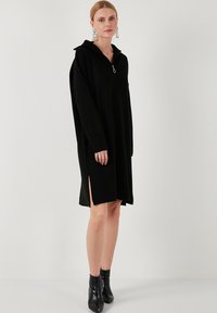 Rochie neagră supradimensionată cu un guler cu fermoar, având slituri laterale și mâneci lungi. Textură netedă și croială lejeră. Botine negre până la gleznă.