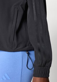 Black athletic jacket featuring three vertical stripes and elastic cuffs. Includes a pocket and an adjustable drawstring at the hem.