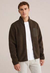 Chaqueta de forro polar marrón con cremallera y alto cuello, superficie texturizada, y un bolsillo con cremallera de un oscuro verde contrastante en el lado izquierdo del pecho. Camiseta blanca debajo.