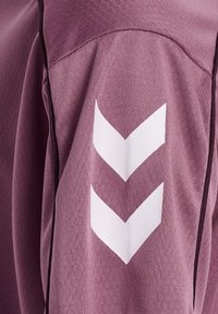Fioletowa koszulka sportowa z teksturowanej tkaniny, udekorowana białym logo w kształcie chevronu na lewym ramieniu i czarnymi wykończeniami wzdłuż szwów.