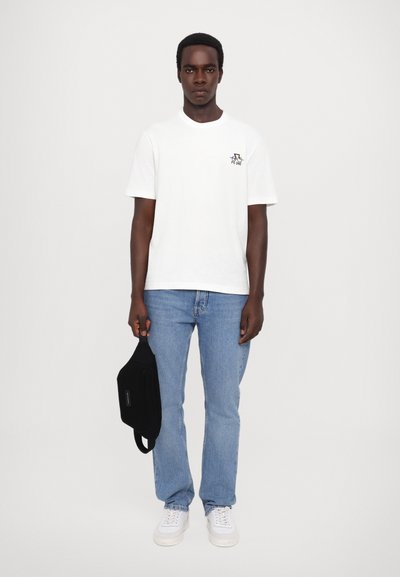 Camiseta de algodón blanca con mangas cortas, que presenta un pequeño logo multicolor en el pecho. Combinada con jeans de mezclilla azul claro y zapatillas blancas.