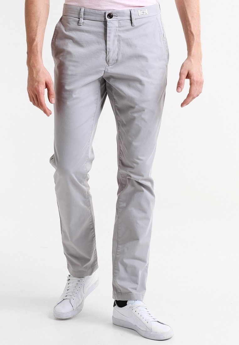 Homme portant un pantalon slim gris clair et des baskets blanches, debout sur un fond blanc uni.