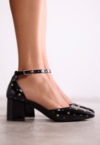 Chaussures en cuir verni noir à talons bloc, ornées de points dorés et argentés, avec une bride à la cheville. Texture lisse avec un design à bout pointu.
