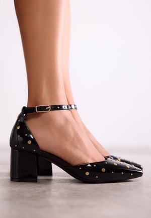 Zapatos de charol negro con tacón cuadrado, adornados con puntos dorados y plateados, y una correa en el tobillo. Textura suave con un diseño de punta afilada.