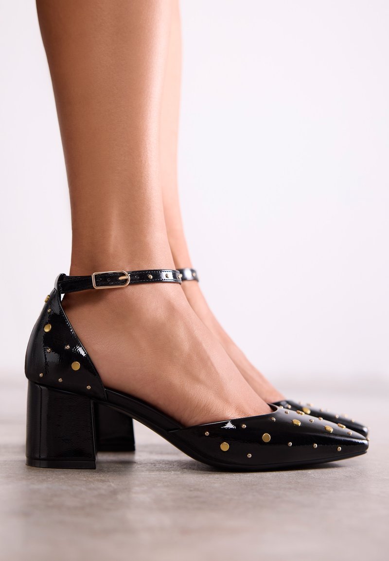 Chaussures en cuir verni noir à talons bloc, ornées de points dorés et argentés, avec une bride à la cheville. Texture lisse avec un design à bout pointu.