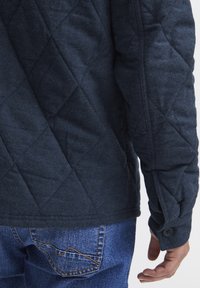 Navy quilted jacket with a textured finish, featuring diamond patterns and buttoned cuffs, paired with blue denim jeans.