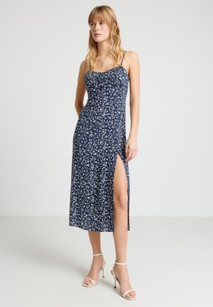 Femme portant une robe midi bleu marine à fleurs avec bretelles fines et fente latérale, associée à des sandales blanches à talons hauts, debout sur un fond blanc.