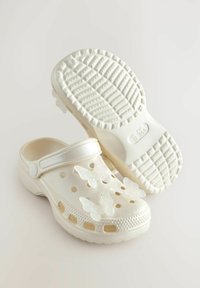 Weiße Clogs mit einer strukturierten Sohle, die über ausgeschnittene Belüftungslöcher und schimmernde Schmetterlingsverzierungen auf der Oberseite verfügen.
