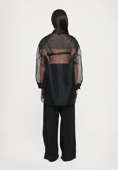 Gjennomsiktig svart bluse med en semitransparent bakside, lange ermer og mansjetter, kombinert med løstsittende svarte bukser. Minimalistisk design, uten mønstre.